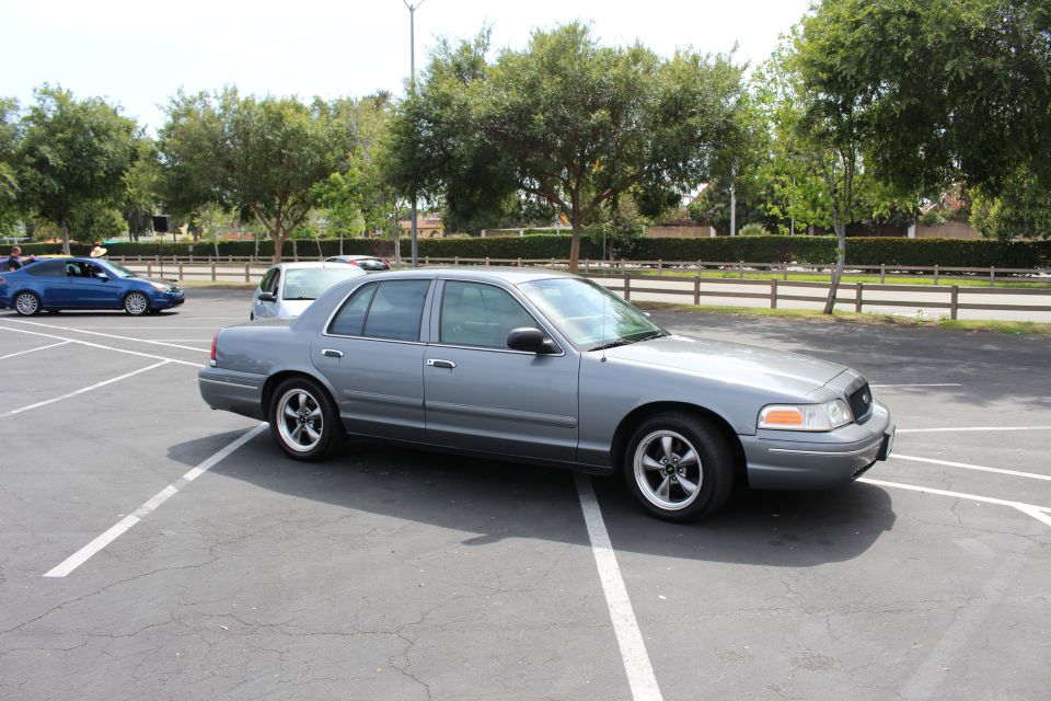 Gray full-size four-door sedan parked in an empty lot with trees and a wooden fence behind