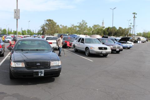 Dark Ford sedan in foreground with rows of cars and a clock tower behind at a car meet