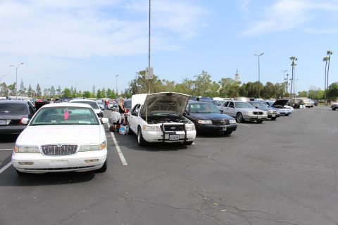 White sedan with raised hood among rows of sedans, distant clock tower under blue sky