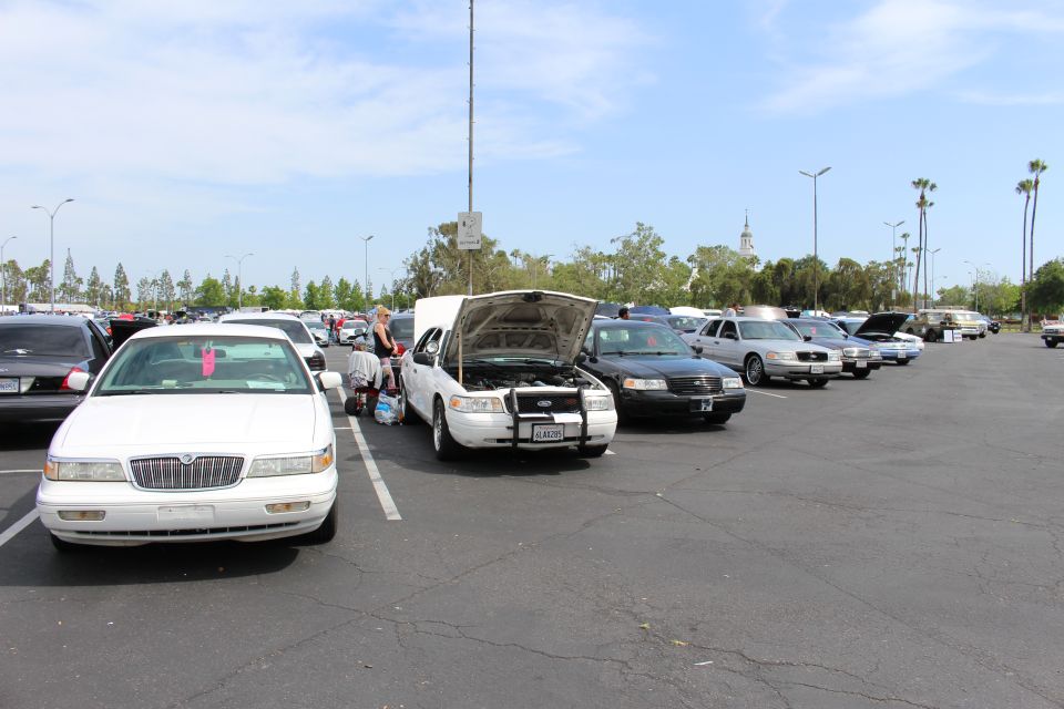 White sedan with raised hood among rows of sedans, distant clock tower under blue sky