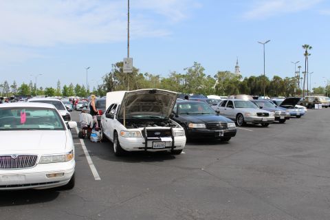 White sedan with open hood and a person beside it, rows of cars stretching back in a lot