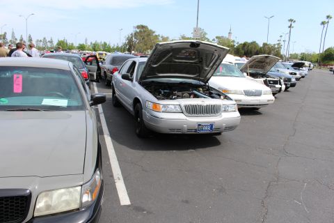 Silver sedan with raised hood in a row of sedans, KAGE plate visible, crowd in background