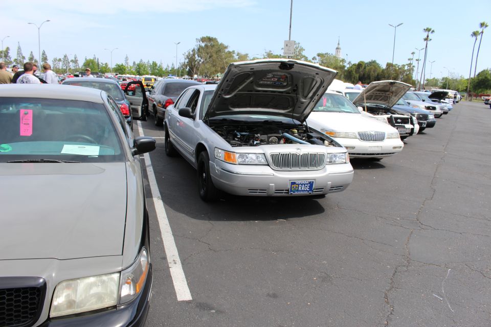 Silver sedan with raised hood in a row of sedans, KAGE plate visible, crowd in background