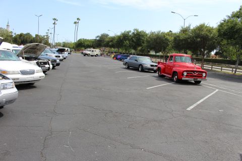 Red classic pickup truck parked at right beside a wide open lot with cars and a fence