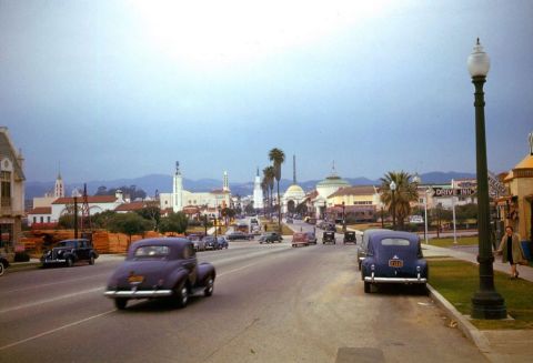 1991 Ford LTD Crown Victoria - A street scene captures multiple vintage vehicles driving down a road lined with buildings and palm trees.