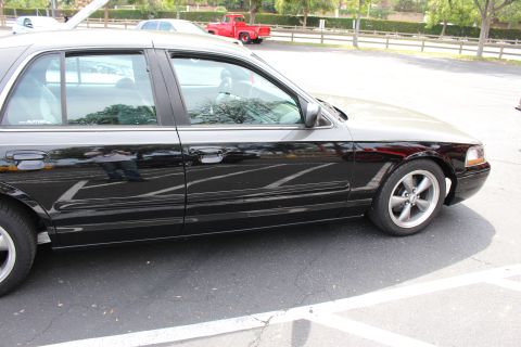 Black full-size sedan in profile with five-spoke wheels, red pickup and fence behind