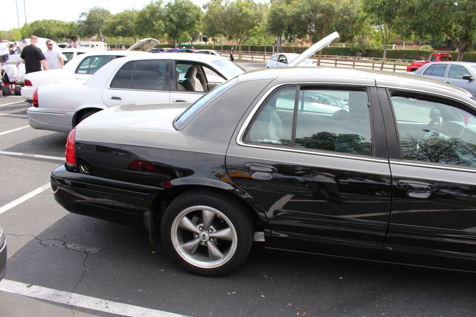 Black sedan rear three-quarter view with five-spoke wheels, white sedan and people behind