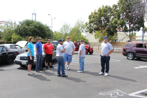 Group of people standing in a parking lot near cars with amusement-park rides behind