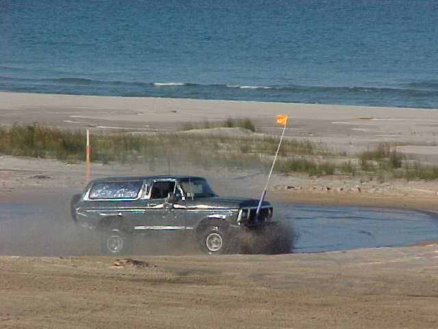 Lifted Ford Bronco with orange whip flag splashing through water on a beach by the ocean