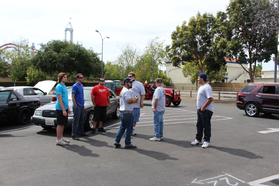 People standing among parked cars with a red vintage vehicle and roller coaster behind