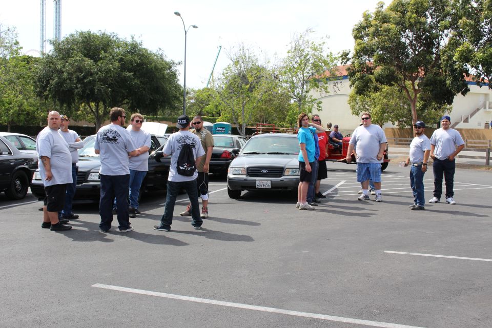 People gathered around parked sedans with a white Ford sedan in the foreground