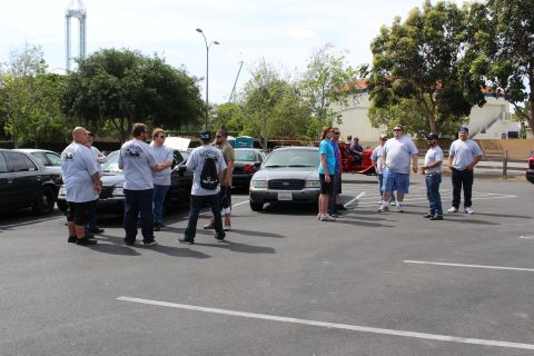 People standing in a parking lot among sedans with a red motorcycle visible