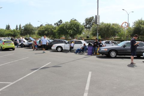 Parking-lot car show with a green hatchback and white sedan, people seated nearby