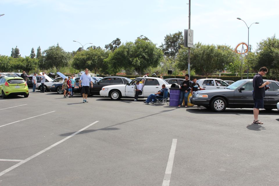 Parking-lot car show with a green hatchback and white sedan, people seated nearby
