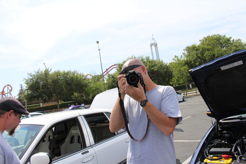 Bald man in gray T-shirt aiming a DSLR camera at a car show, amusement-park rides behind