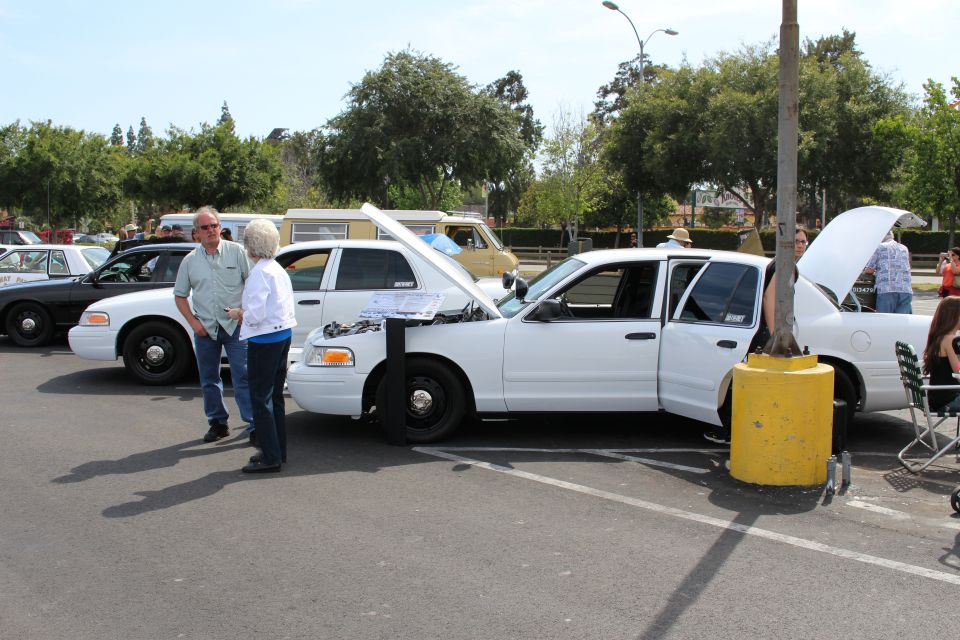 White Ford Crown Victoria with open hood beside other sedans at a car show