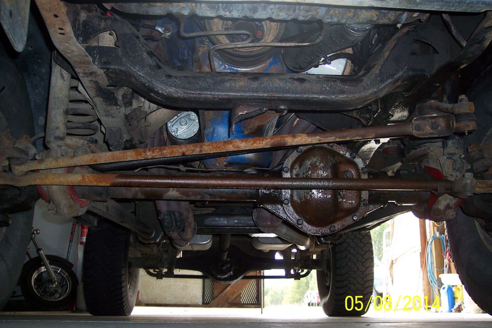 Underside view of a vehicle's suspension and drivetrain components. Shows rusty metal parts and a tire