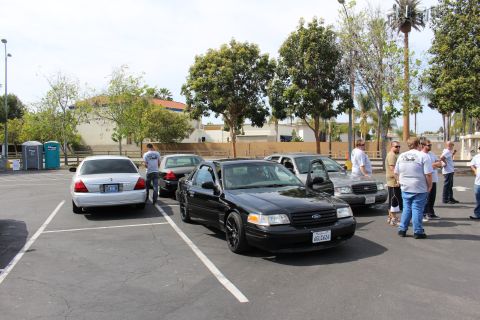 Black Ford sedan with black wheels parked among cars, people gathered nearby, portable toilet behind