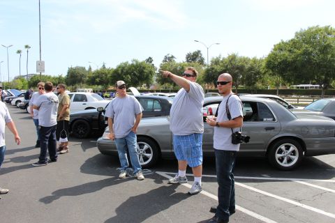 Men standing in parking lot beside silver sedans, one pointing, another holding a camera
