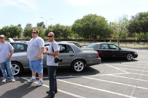 Men standing beside silver and black sedans in a parking lot under bright sun