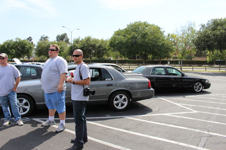 Men standing beside silver and black sedans in a parking lot under bright sun