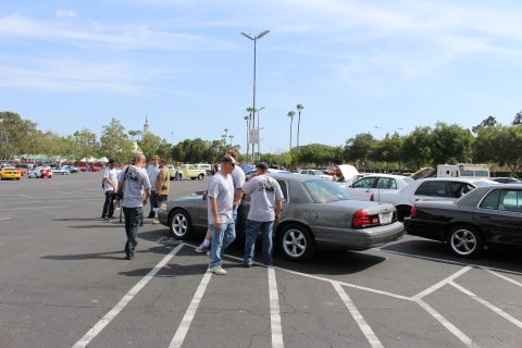 People walking among silver sedans in a large parking lot car meet with palm trees