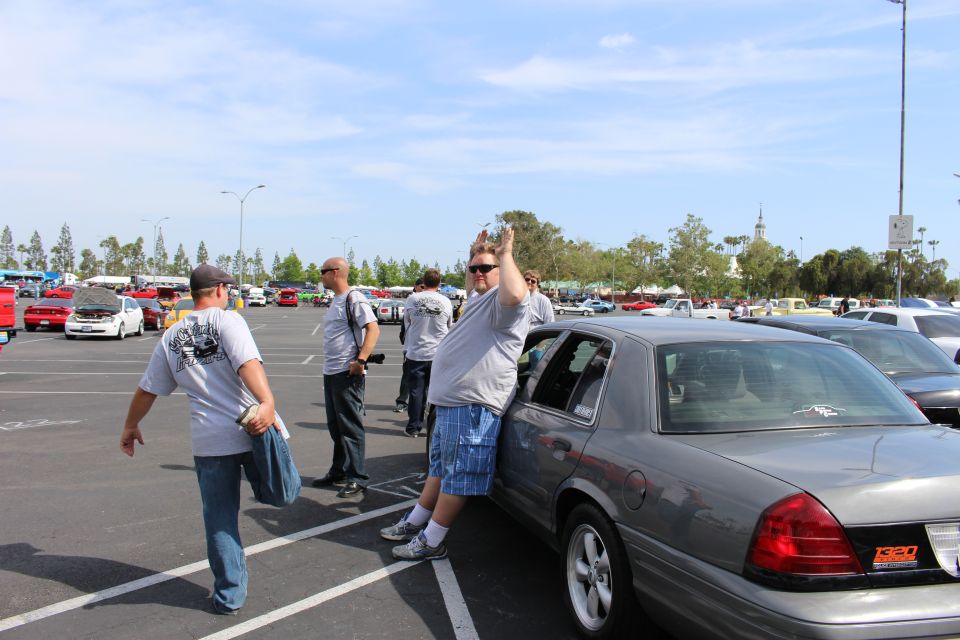 Man leaning on silver sedan waving as others walk past in a large car-meet parking lot