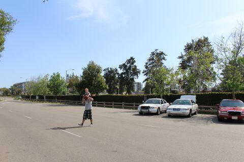 Man holding a child walks across a lot near parked sedans and trees