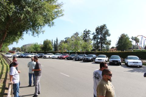 People standing and talking in a lot with parked cars and a roller coaster behind