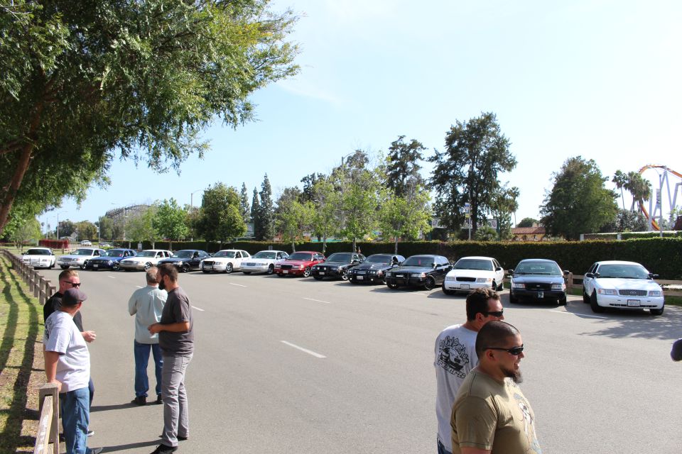 People standing and talking in a lot with parked cars and a roller coaster behind