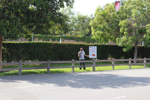 Man standing by a roadside sign in a parking lot with trees and a fence