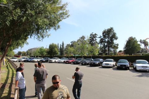 People and parked cars in a lot at a car meet with trees behind