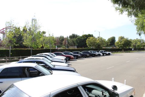 Wide parking lot with a row of cars along a hedge and a tower ride behind