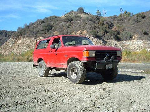 1988 Ford Bronco - This red 1988 Ford Bronco is parked on a dirt road with a hilly landscape behind it.