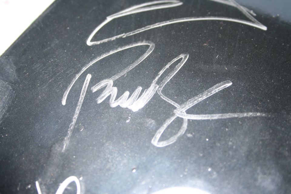 Close-up of a white autograph on a dark, possibly metallic surface. Text from a website is visible at the bottom right