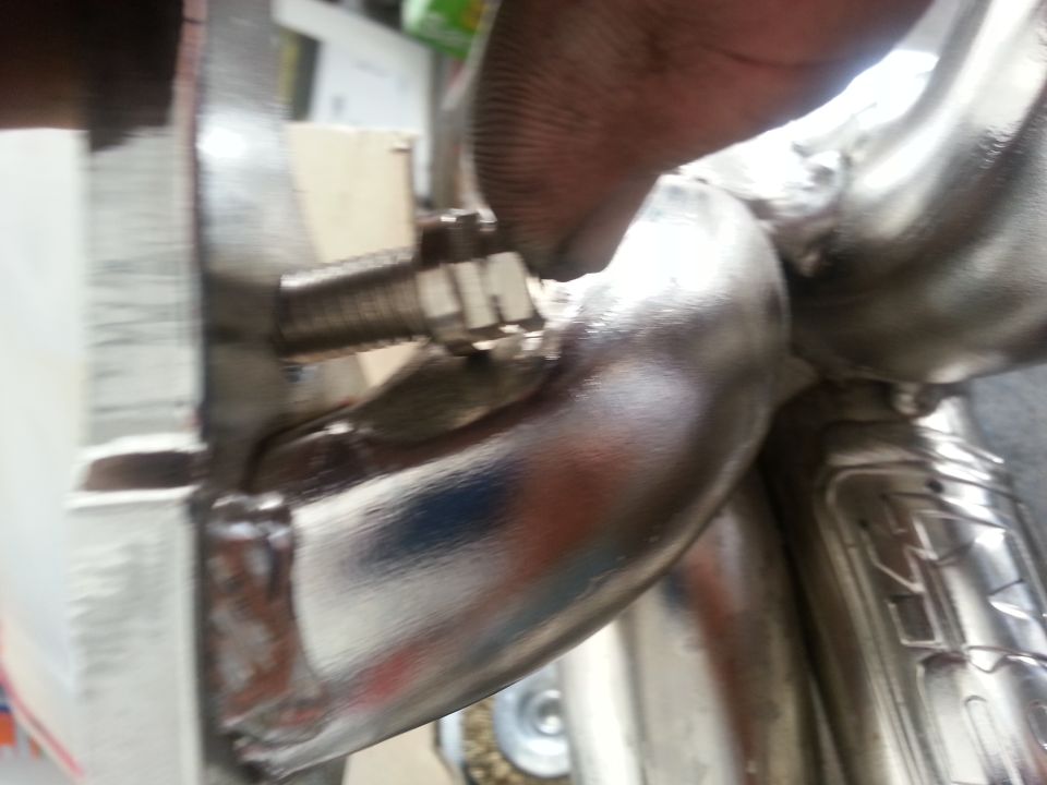 Close-up of a shiny metal exhaust manifold with a bolt and nut. Text 'SUPERMOTORS www.supermotors.net' is visible