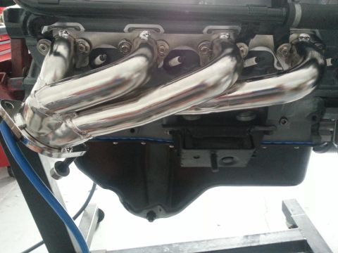 1994 Ford Bronco - Close-up of a shiny exhaust manifold attached to an engine, with a visible website logo at the bottom.