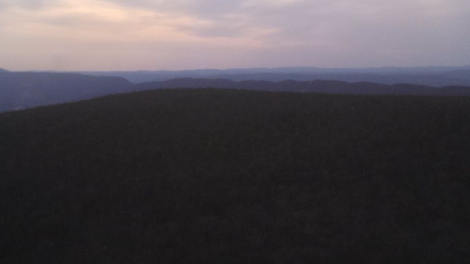 Dark, forested hills under a pale, cloudy sky with a Supermotors logo in the lower right corner