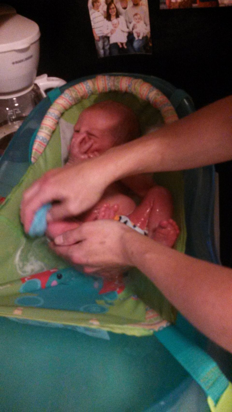 Baby in a bath seat with hands washing it. Family photo on the wall in background