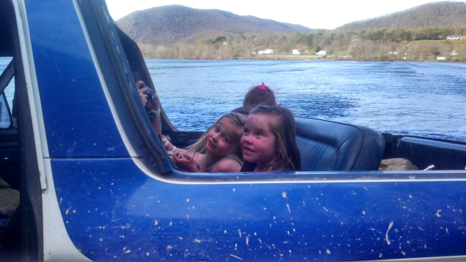 Three children look out from the interior of a blue vehicle, with a body of water and hills in the background
