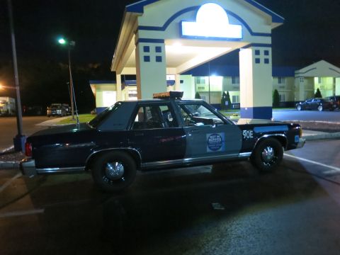 1991 Ford LTD Crown Victoria - blue State Police sedan marked 90E with a Not In Service placard sits at night under a lit motel portico.