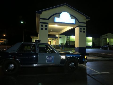 1991 Ford LTD Crown Victoria - Side view of a dark State Police sedan marked 90E with a Not In Service placard parked at night beside a lit motel.