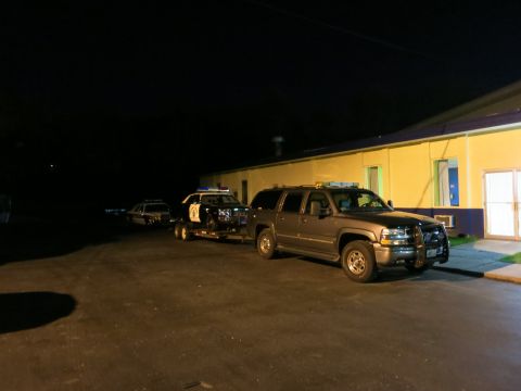 1991 Ford LTD Crown Victoria - dark SUV tows a trailer carrying a police car at night in a lot beside a yellow building, another car nearby.