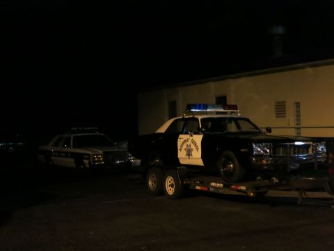 1991 Ford LTD Crown Victoria - black-and-white Highway Patrol sedan sits on a flatbed trailer at night beside a building, another cruiser at left.