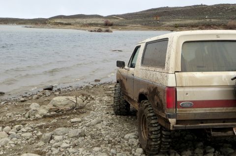 1989 Ford Bronco - A muddy 1989 Ford Bronco is parked on a rocky shore next to a body of water.