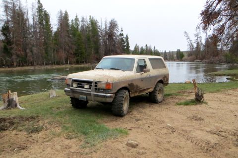 1989 Ford Bronco - Muddy 1989 Ford Bronco sits by a river with trees in the background.