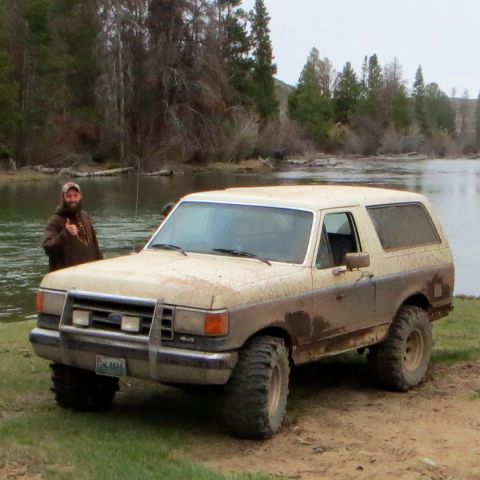 1989 Ford Bronco - A man stands next to a dirty Ford Bronco near a body of water with trees in the background.