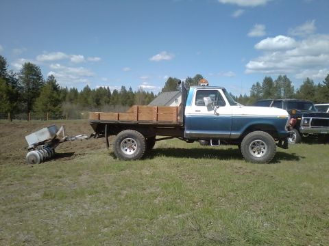 1973 Ford F-250 - A blue and white flatbed truck is parked in a grassy field, hitched to a tiller attachment.