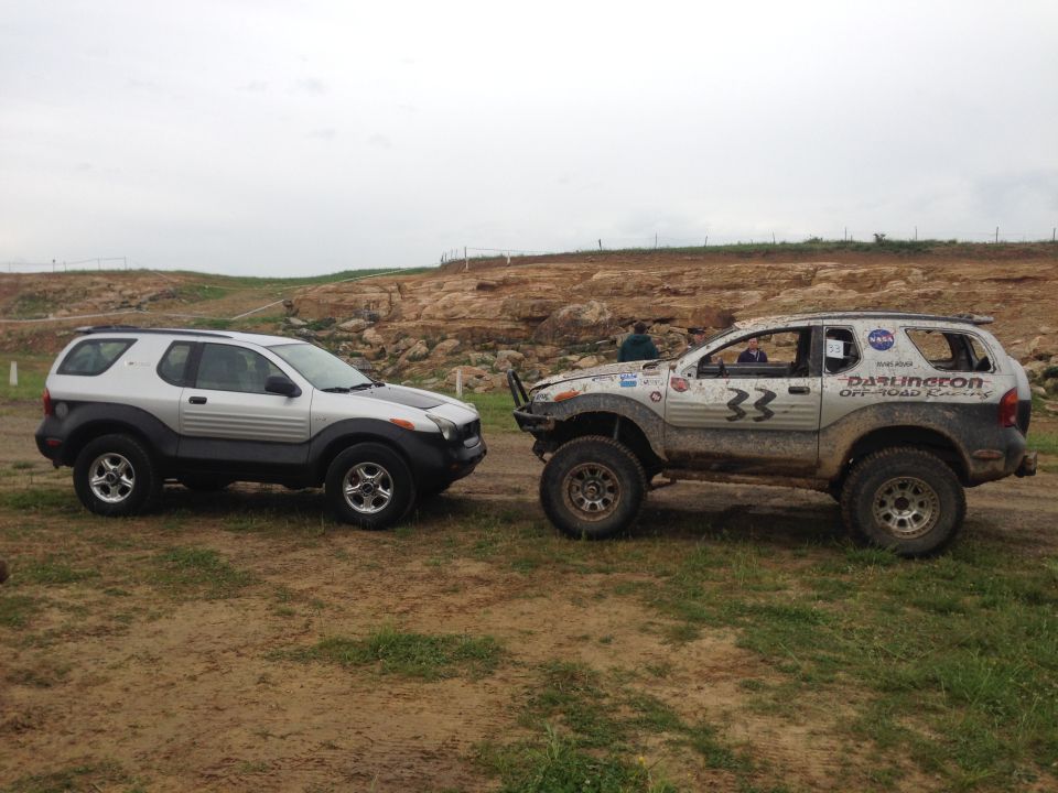 Two 1999 Isuzu Vehicross SUVs parked side-by-side on a dirt track with a rocky hill in the background