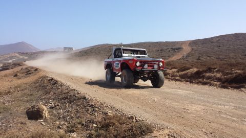1996 Ford Bronco - A red and white Ford Bronco races down a dusty off-road track under a clear blue sky.